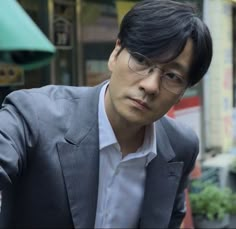 a man wearing glasses and a suit is standing in front of a store with his hand on his hip