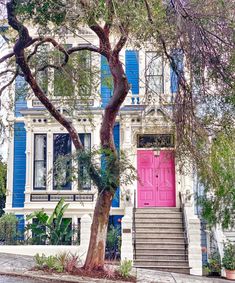 a pink door is in front of a white house with blue shutters and trees