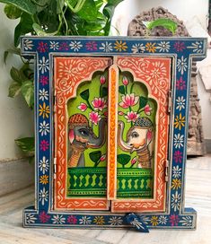 an intricately painted wooden box with two elephants and flowers on the front, sitting next to a potted plant