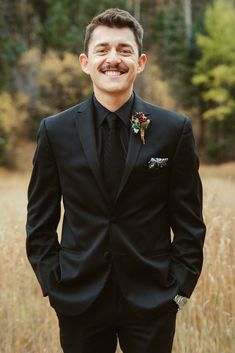 a man in a black suit and flowered boutonniere smiles at the camera