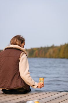 a woman sitting on a dock holding a can of beer and looking at the water