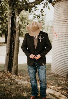 a man wearing a cowboy hat standing in front of a tree with his hands on his hips