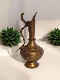 two brass vases sitting next to each other on a white surface with succulent plants in the background