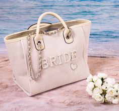 a white purse sitting on top of a sandy beach next to a bouquet of flowers