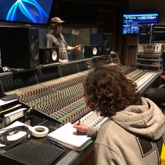 two people sitting at a mixing desk in front of some monitors and recording equipment with one person standing behind the sound board