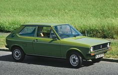 an old green car is parked on the side of the road in front of some tall grass