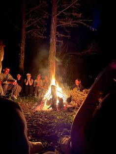 a group of people sitting around a fire in the woods at night with lights on