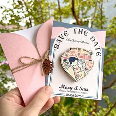 someone holding up a save the date card with a heart shaped pin attached to it