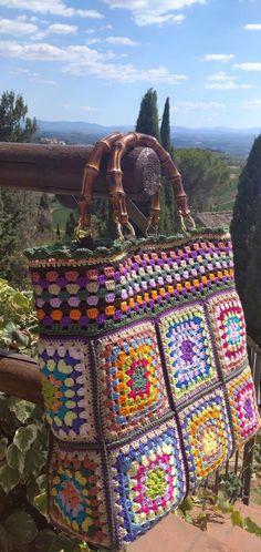 a multicolored crocheted bag sitting on top of a wooden rail next to trees