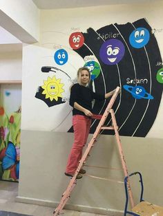 a woman standing on a ladder in front of a wall with cartoon faces painted on it