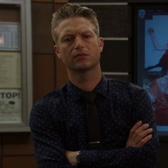 a man with his arms crossed standing in front of a tv screen wearing a shirt and tie