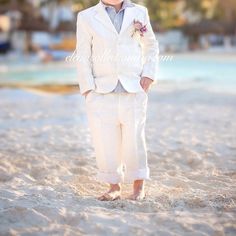 a little boy in a white suit and tie standing on the beach with his hands in his pockets