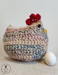 a crocheted chicken sitting next to an egg on a table with a white wall in the background