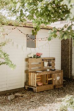 a wooden table sitting under a tree next to a white building with flowers on it