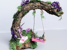 a miniature tree swing with purple flowers and moss on the top, hanging from a branch