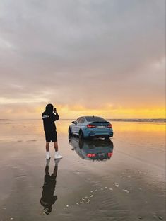 a person standing in front of a car on the beach with water reflecting off it's surface