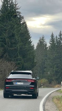 the rear end of a black car driving on a road near some trees and bushes