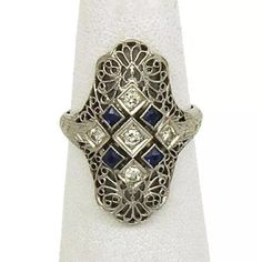 This is a beautiful Art Deco ring, it is crafted from 18k white with milgrain and filigree. The entire long length of this ring has a lovely floral filigree design with round cut diamonds in small square shape form and bezel set square cut sapphires. It has the metal content stamp.    Material: 18k white gold  Hallmark:  18k  Measurement: 23.8mm long x 17.5mm across x 4mm high  Gemstone:  Square cut sapphire  Diamond: round cut   Ring Size: 6  Weight: 4.1 grams Art Deco Formal Filigree Ring With Gemstone, Art Deco Filigree Ring With Center Stone, Blue Filigree Art Deco Jewelry, Art Deco Silver Filigree Ring Hallmarked, Art Deco Diamond Filigree Ring Hallmarked, Round Cut Ring, Filigree Ring Gold, Floral Filigree, Deco Ring