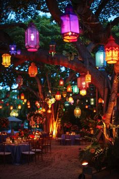 many colorful lanterns are hanging from the tree in this outdoor dinner area with tables and chairs