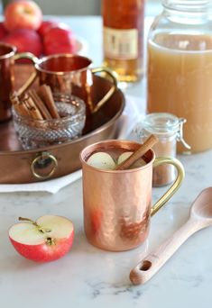 an apple cider, cinnamon sticks and apples on a marble table with copper mugs