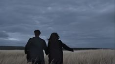 two people are walking through the tall grass in front of an overcast sky at night