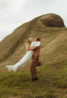 a man carrying a woman in his arms on top of a grass covered hill with a large rock behind him