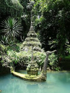 a statue in the middle of a river surrounded by trees and bushes with moss growing on it's sides