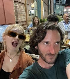 two people taking a selfie in front of a group of people at an outdoor restaurant