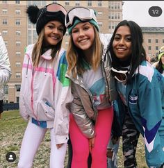 three girls in ski gear posing for the camera with snowboarders and buildings in the background