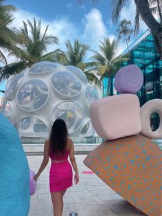 a woman in a pink dress is walking by some large sculptures and palm trees on the beach