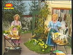 two women are standing in front of a house with flowers and gardening equipment on the porch