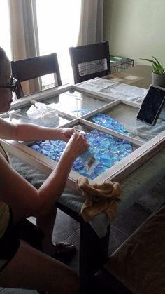 a woman sitting at a table working on an art project with blue glass and wood