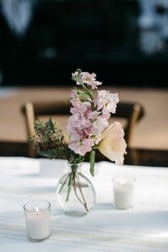 a vase with flowers and candles on a white table cloth next to two wooden chairs