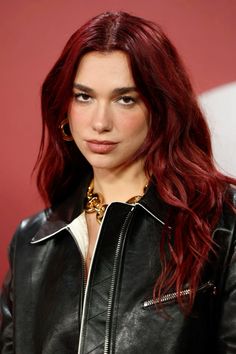 a woman with red hair wearing a black leather jacket and gold jewelry, standing in front of a red wall