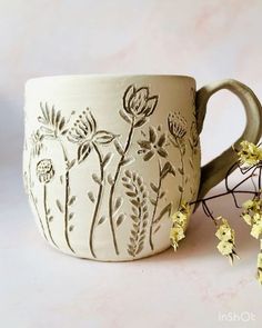 a mug with flowers painted on it next to some yellow flowers and leaves in the background