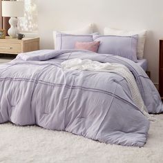 a bed with purple comforter and pillows in a white room next to a dresser