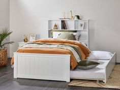 a white bed with an orange blanket on top of it next to a book shelf