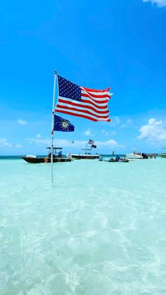 an american flag flying in the ocean next to boats on the beach with blue sky