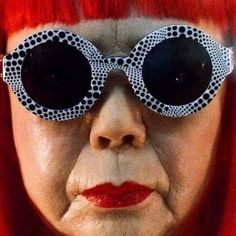 an old woman with red hair and big round glasses on her face is staring at the camera