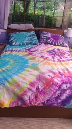 a bed covered in a colorful tie - dyed comforter next to a window with curtains