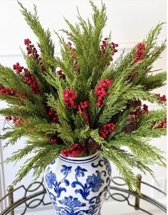 a blue and white vase with red berries on it sitting on a metal table next to a pine cone