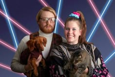 a man and woman holding two cats in front of a colorful background with neon lights