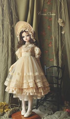 the doll is wearing a dress and hat with flowers on it's head, while standing in front of a chair