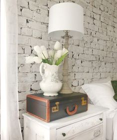 a white vase with flowers sitting on top of an old suitcase next to a bed