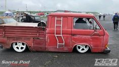 an old red truck parked in a parking lot next to other cars and people walking around