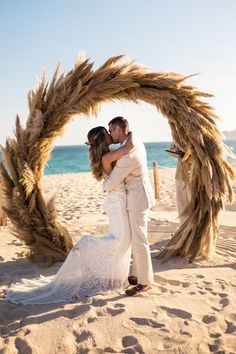 a bride and groom are standing under an arch made out of dried grass on the beach