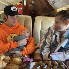 a man holding a baby while sitting next to a woman and tray of doughnuts