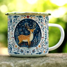 a white and blue mug with a deer on it sitting on a wooden table in front of some trees