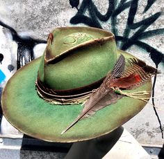 LADY JANE is a transformed Olive & Pique Kaia hat. It's structured, with a flat brim, and a 4.25" crown rise. The brim is 3 1/4 and the head size is adjustable up to 23" It's been reshaped, dyed with various shades of green, burned, stitched and etched with a paisley & floral design.  The band is cowhide, overlaid with bandana, leather cord, and twine.  It's tied off with a large turkey feather, a Guinea hen feather and an African free trade bead. Please feel free to ask questions. Free Trade, Boho Hat, Festival Hat, Happy Fourth Of July, Hat Ideas, Paisley Floral, Trade Beads, Cowgirl Style, Custom Hats