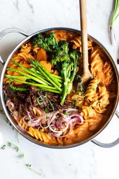 a pot filled with pasta and vegetables on top of a white counter next to a wooden spoon